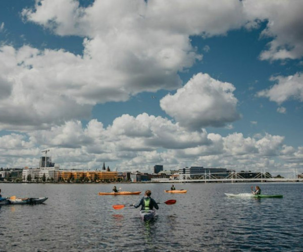 Stadssiluett och bl&aring; himmel med vita moln och en sj&ouml; i f&ouml;rgrunden d&auml;r n&aring;gra paddlar kajak.